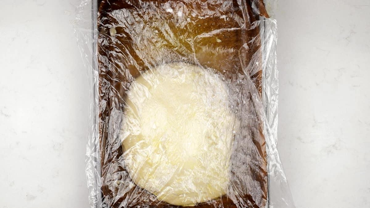 Round ball of dough for sourdough focaccia rising on a brown tray, covered with clear plastic wrap, on a white countertop.