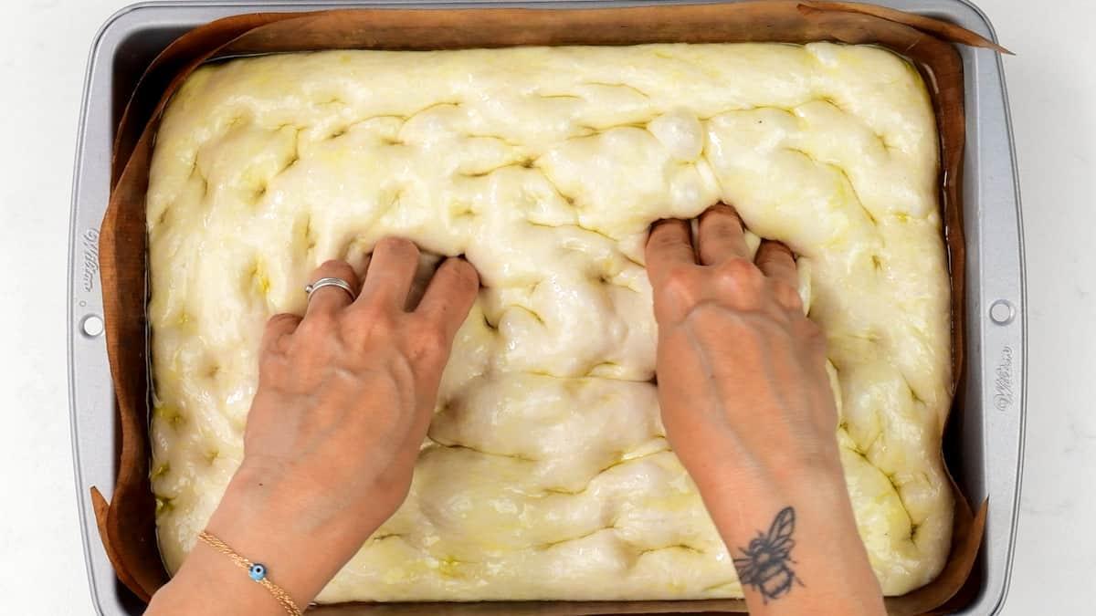 Two hands press dimples into uncooked sourdough focaccia dough in a parchment-lined baking pan. The persons left wrist shows a bracelet and a butterfly tattoo—perfect for anyone trying an easy sourdough focaccia recipe.