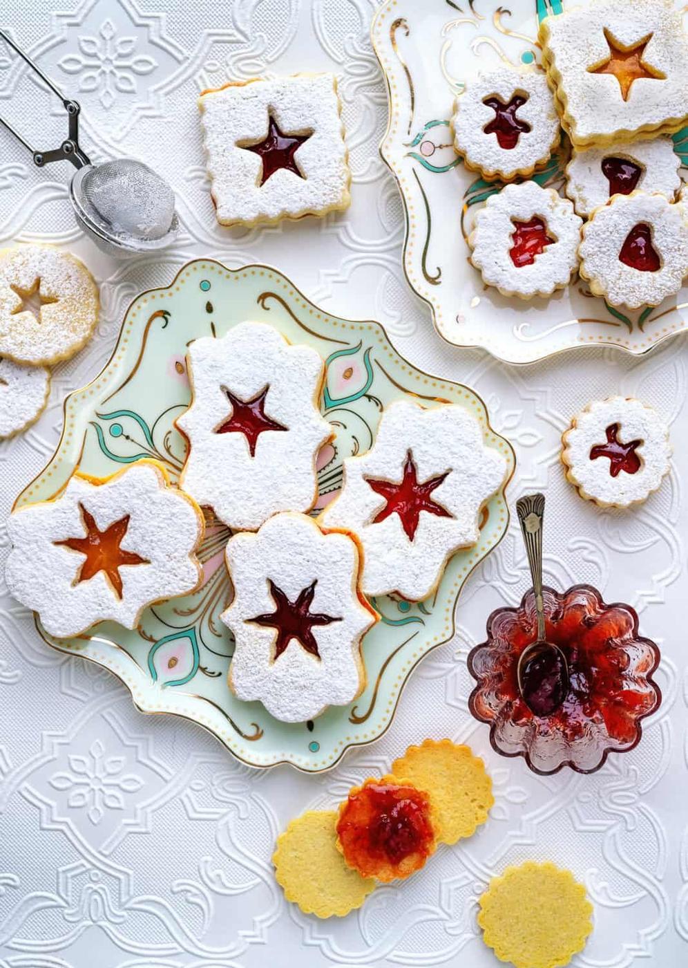 Assorted Austrian Linzer cookies with powdered sugar and jam filling are arranged on decorative plates, alongside a jar of jam and a sifter of powdered sugar on a white embroidered tablecloth.