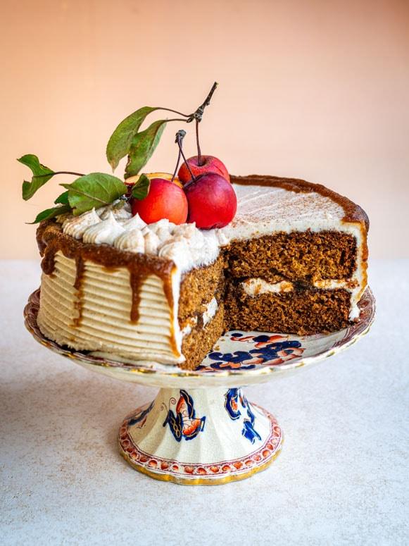 A spiced apple butter cake with white frosting and caramel drizzle sits on a decorative cake stand. Topped with whipped cream, leaves, and three red plums, this Thanksgiving dessert has a large slice cut to reveal its layers.