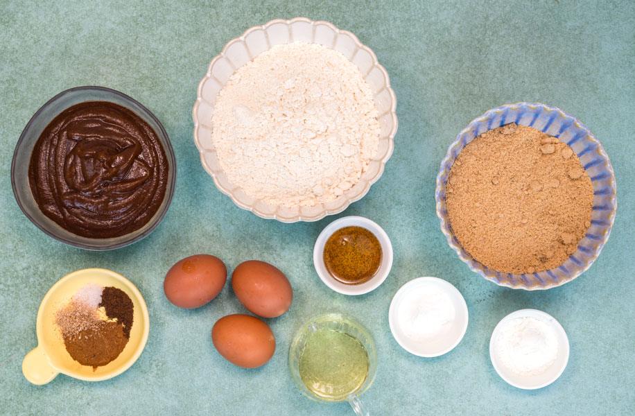 A top-down view of baking ingredients in bowls and cups on a teal surface, including flour, brown sugar, chocolate batter, spices, three eggs, oil, and white powders.