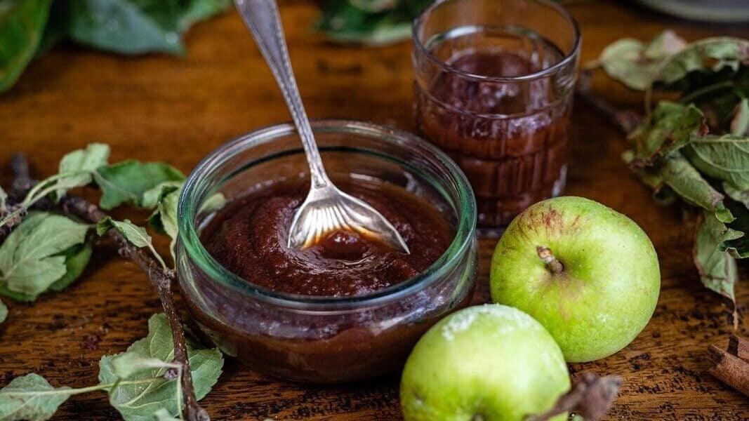 A glass bowl of apple butter with a spoon, a glass of juice, green apples, and apple branches on a wooden table, with a bowl of apples in the background.