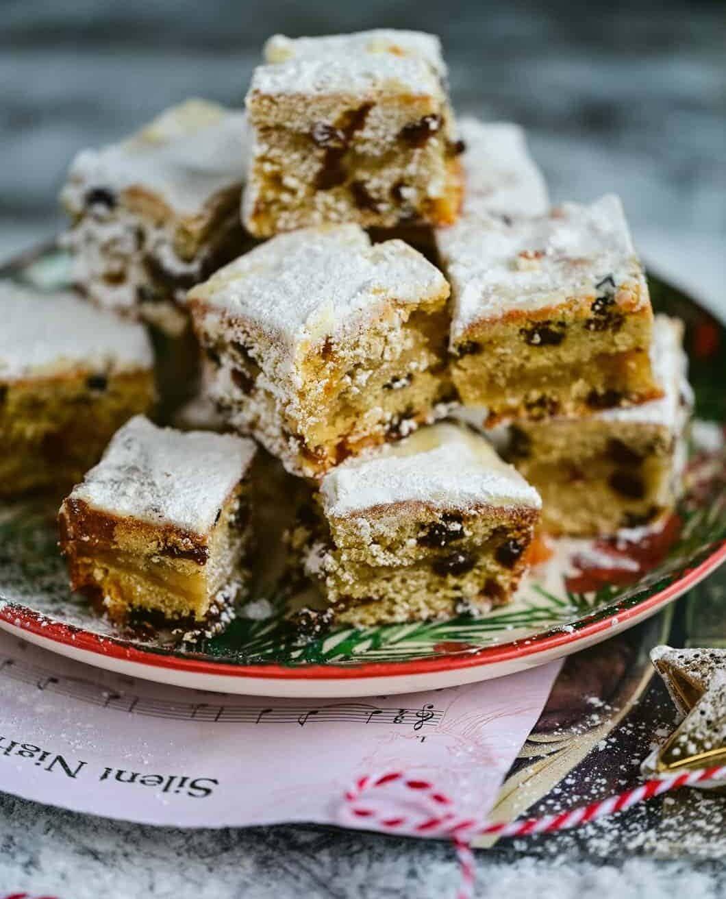 A plate stacked with golden-brown Stollen Bites, dusted with powdered sugar and dotted with currants or raisins. The festive treats rest on colorful papers and sheet music, ready to enjoy.