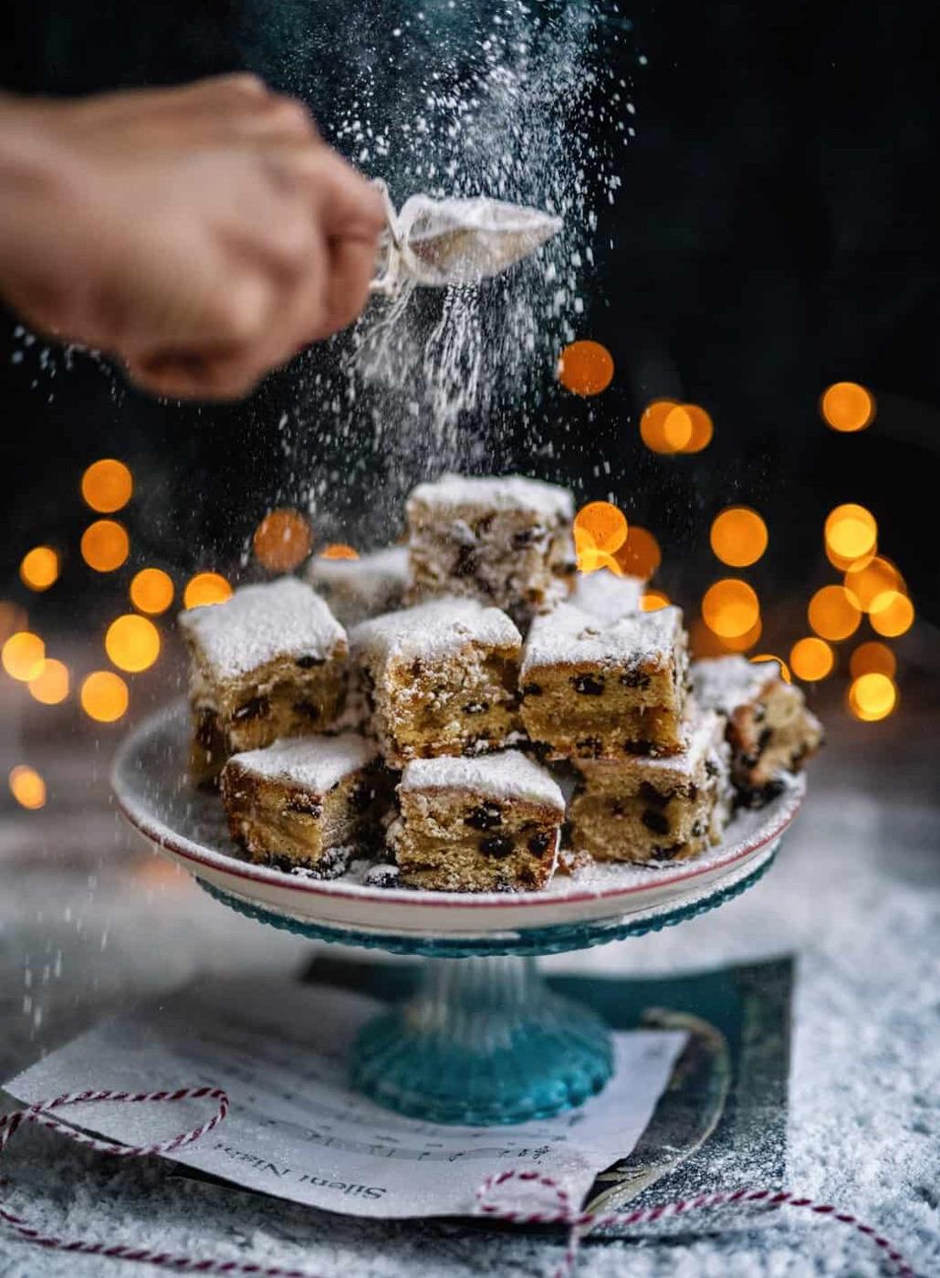 A hand sprinkles powdered sugar over a stack of German cake squares with fruit on a decorative cake stand. Warm, blurred lights glow in the background, creating a festive, cozy Christmas atmosphere—no yeast required.