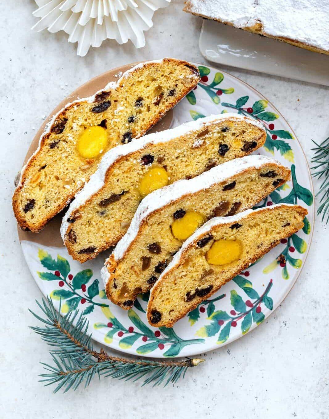 Four slices of fruit-studded stollen cake dusted with powdered sugar are arranged on a decorative plate with green and red holiday patterns, along with a pine twig as a festive accent.