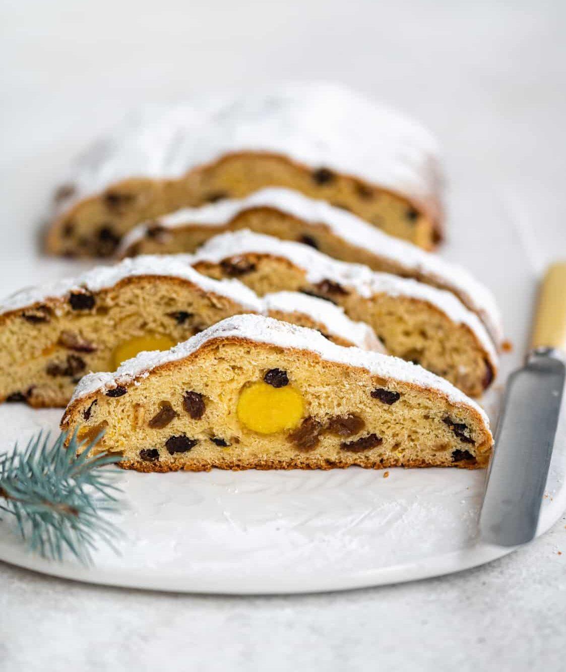 Sliced loaf of traditional German Stollen with raisins and a yellow marzipan center, dusted with powdered sugar, on a white plate next to a knife and a small pine twig—a classic stollen cake perfect for the holidays.