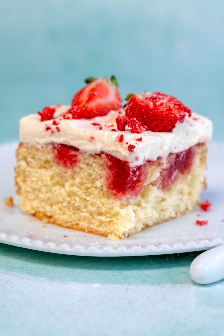 A slice of vanilla cake topped with white frosting, strawberry sauce, and fresh strawberries sits on a white plate against a light blue background.