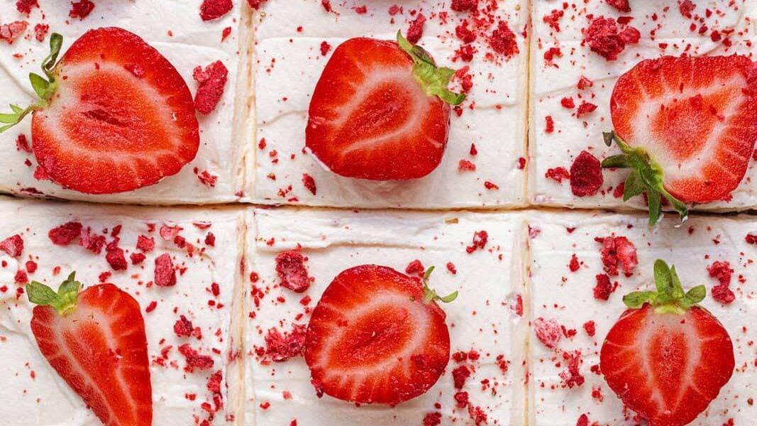 Overhead view of sliced cake squares topped with halved fresh strawberries and sprinkled with crushed dried strawberries on white frosting.