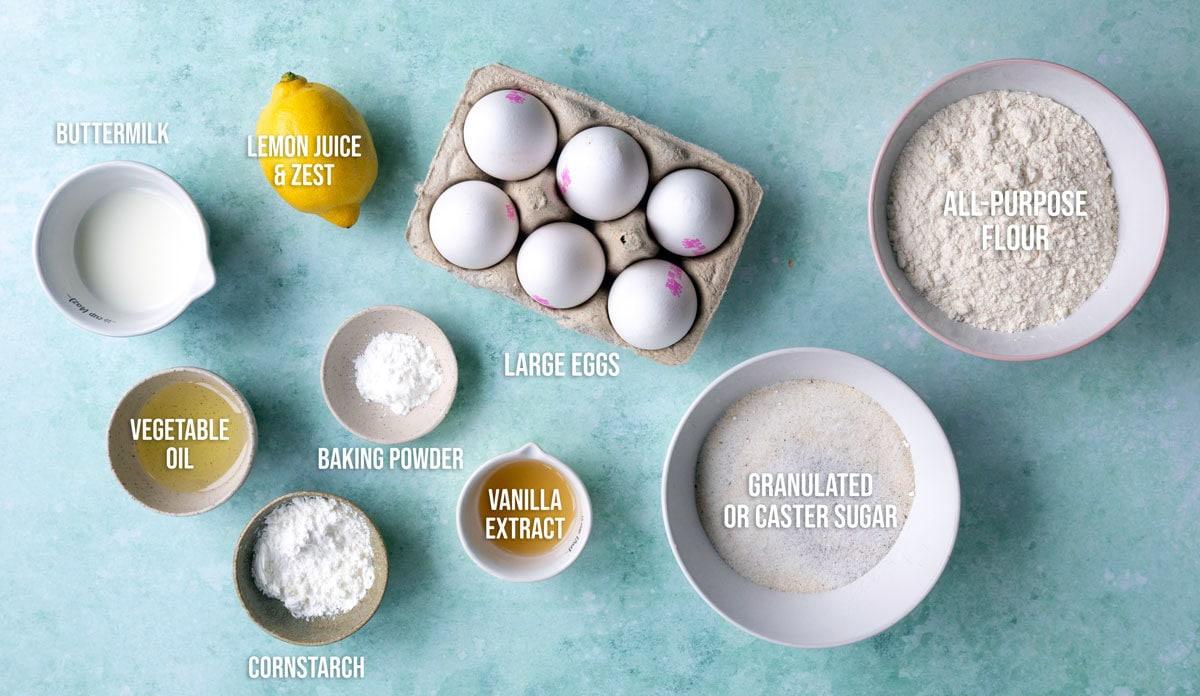 Top-down view of labeled baking ingredients on a blue surface, including buttermilk, lemon, eggs, flour, sugar, oil, baking powder, cornstarch, and vanilla extract, arranged in bowls and containers.