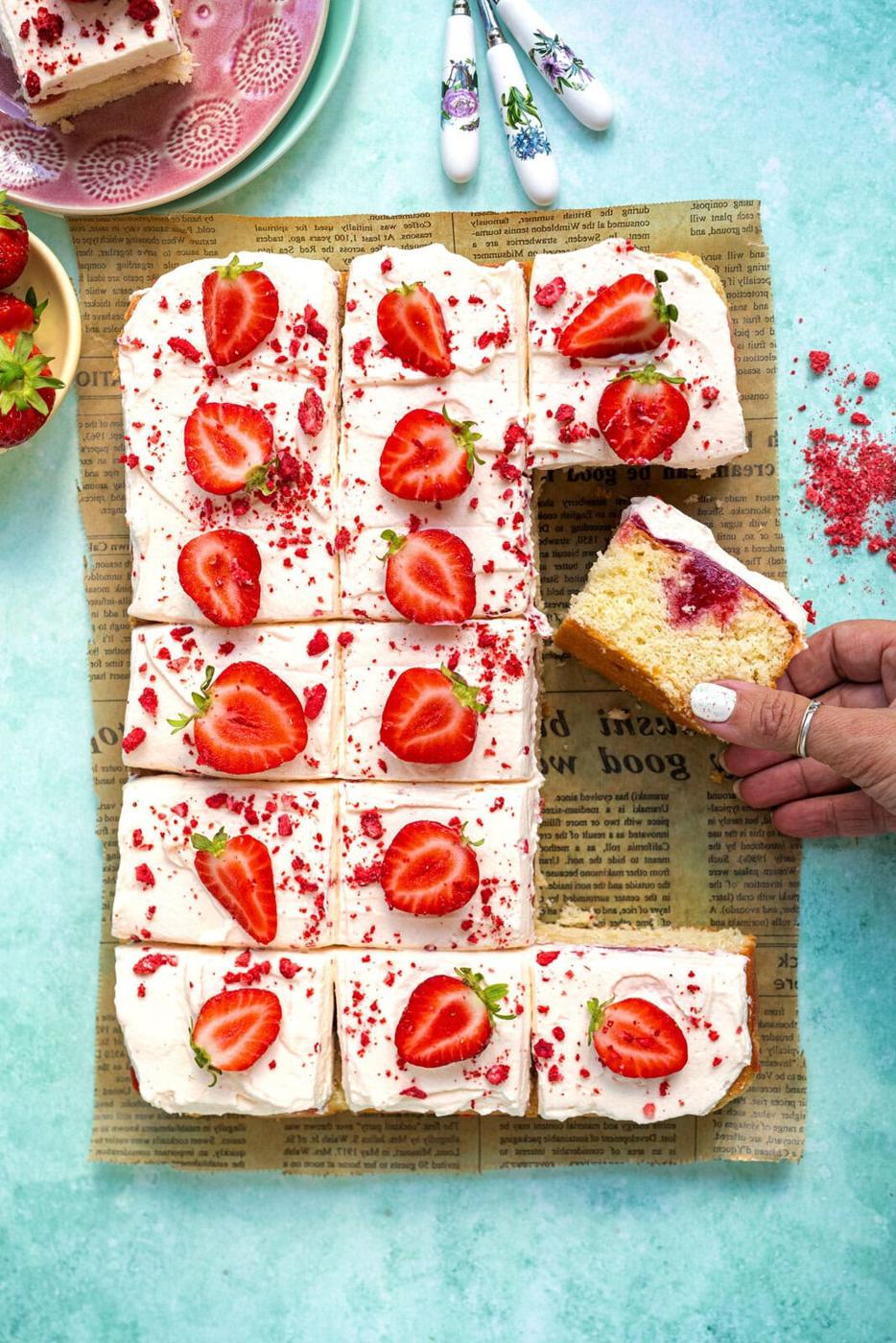 A tray of sliced vanilla cake with white frosting, topped with halved fresh strawberries and red sprinkles, sits on newspaper. A hand is lifting one piece, revealing a layer of strawberry jam inside.