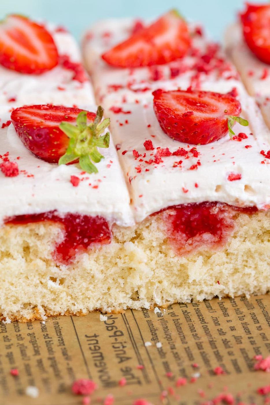 A close-up of sliced vanilla cake topped with white frosting, halved fresh strawberries, and red sprinkles, set on a piece of printed parchment paper. The cake appears fluffy and moist with strawberry filling inside.