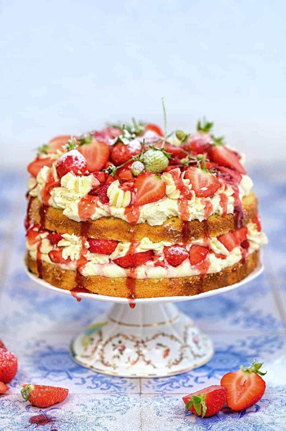 A stunning Strawberry Shortcake Cake sits on a decorative stand, its two layers filled and topped with cream, fresh strawberries, and strawberry sauce, surrounded by whole and sliced berries on a blue patterned surface.