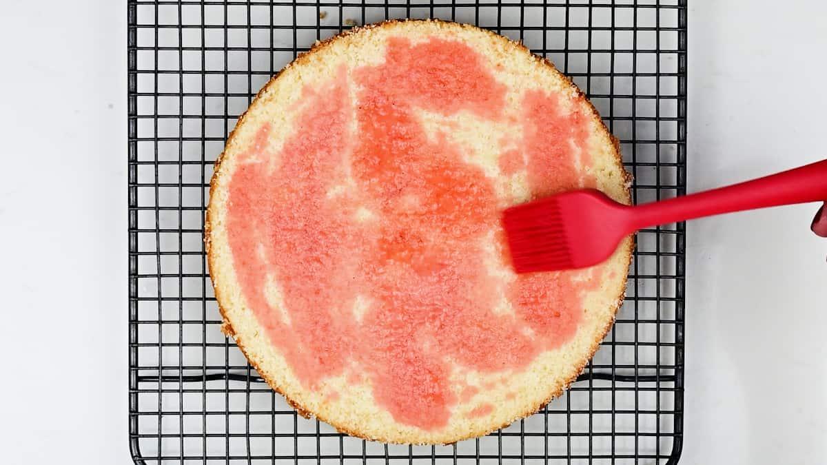A round Strawberry Shortcake Cake sits on a cooling rack while a red silicone brush spreads a glossy pink liquid over the cake’s surface.