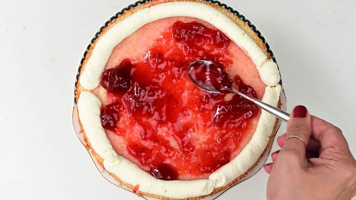 A hand spreads bright red jam over a Strawberry Shortcake Cake topped with a ring of white whipped cream, using a spoon. The cake sits on a white surface.