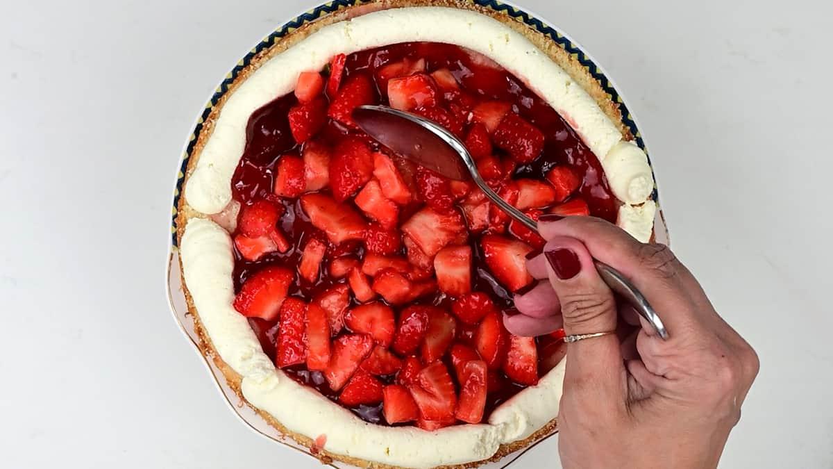 A hand holding a spoon is spreading a layer of fresh strawberry topping over a cheesecake with a cream border, creating the look of a Strawberry Shortcake Cake, seen from above on a white background.