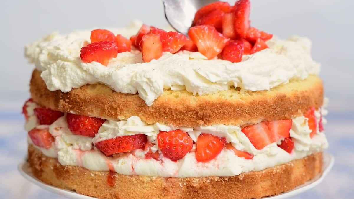 A classic Strawberry Shortcake Cake, featuring a light sponge cake layered with whipped cream and sliced strawberries, then topped with extra whipped cream and fresh strawberries as a spoon adds the finishing touch.