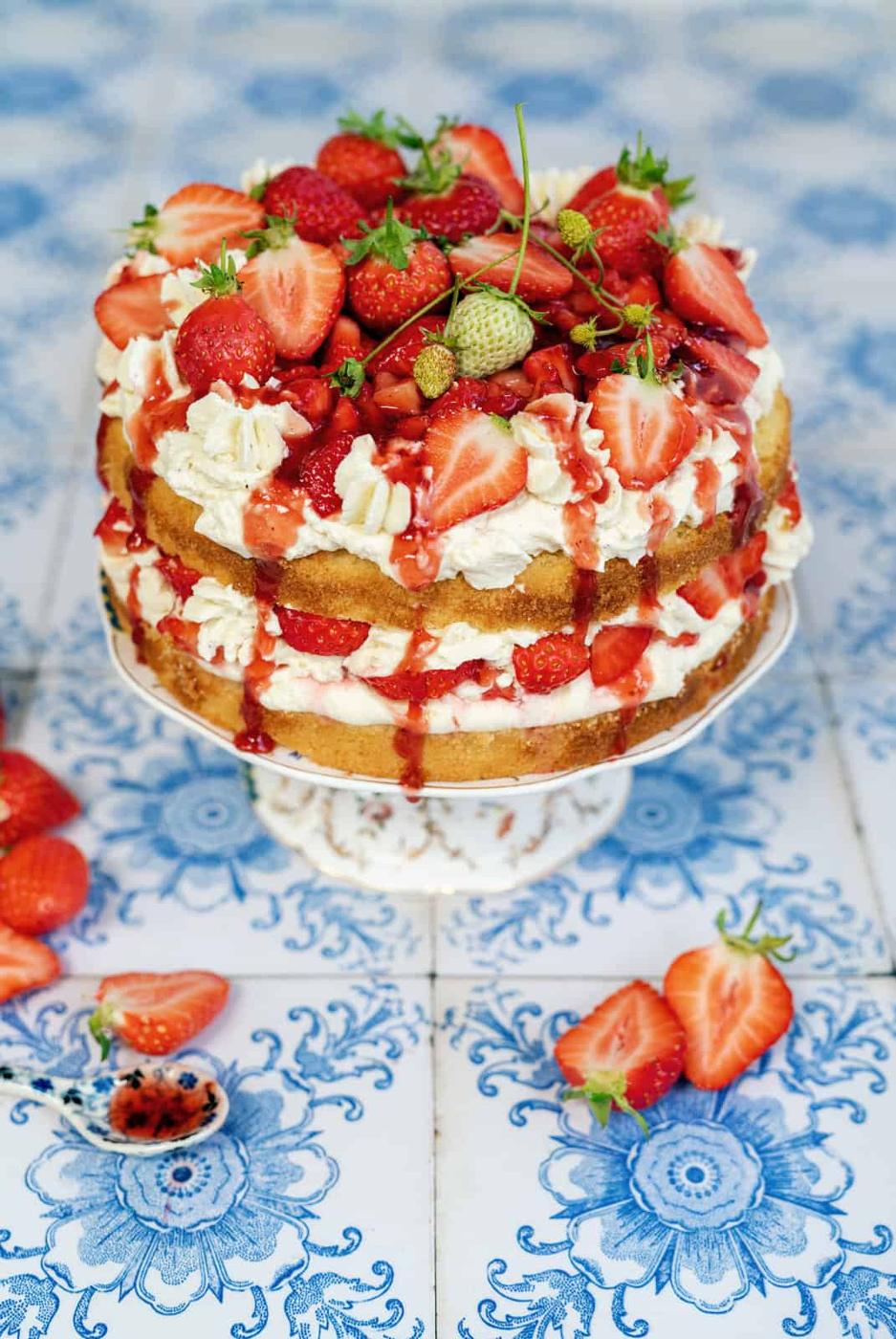 A three-layer Strawberry Shortcake Cake filled with whipped cream and topped with fresh strawberries and strawberry sauce, displayed on a cake stand with strawberries scattered around on a blue and white patterned surface.