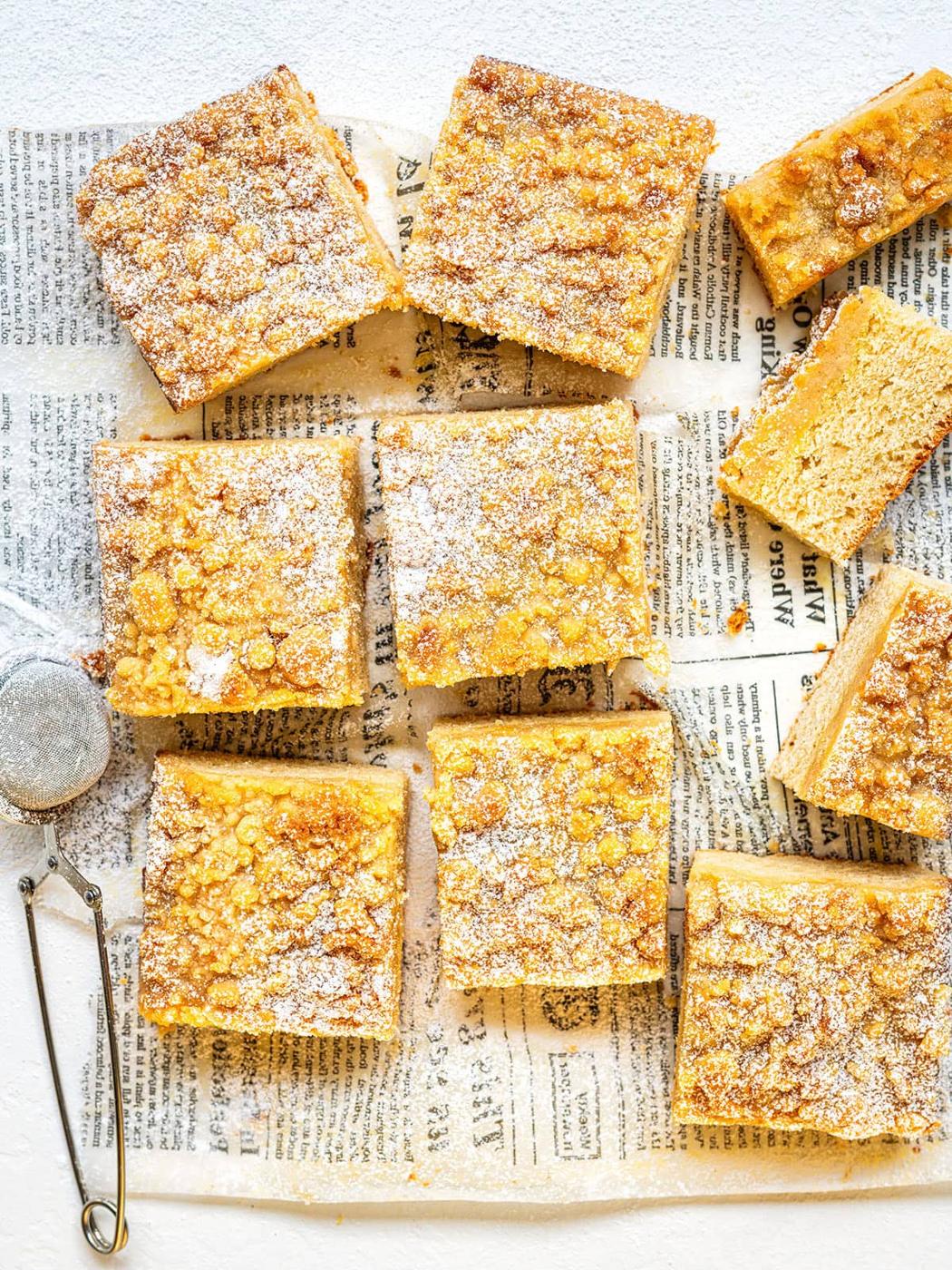 Nine squares of German crumb cake, or streuselkuchen, topped with powdered sugar rest on newspaper print parchment; a tea strainer sits nearby, and one piece is partly eaten, revealing its soft, layered interior.