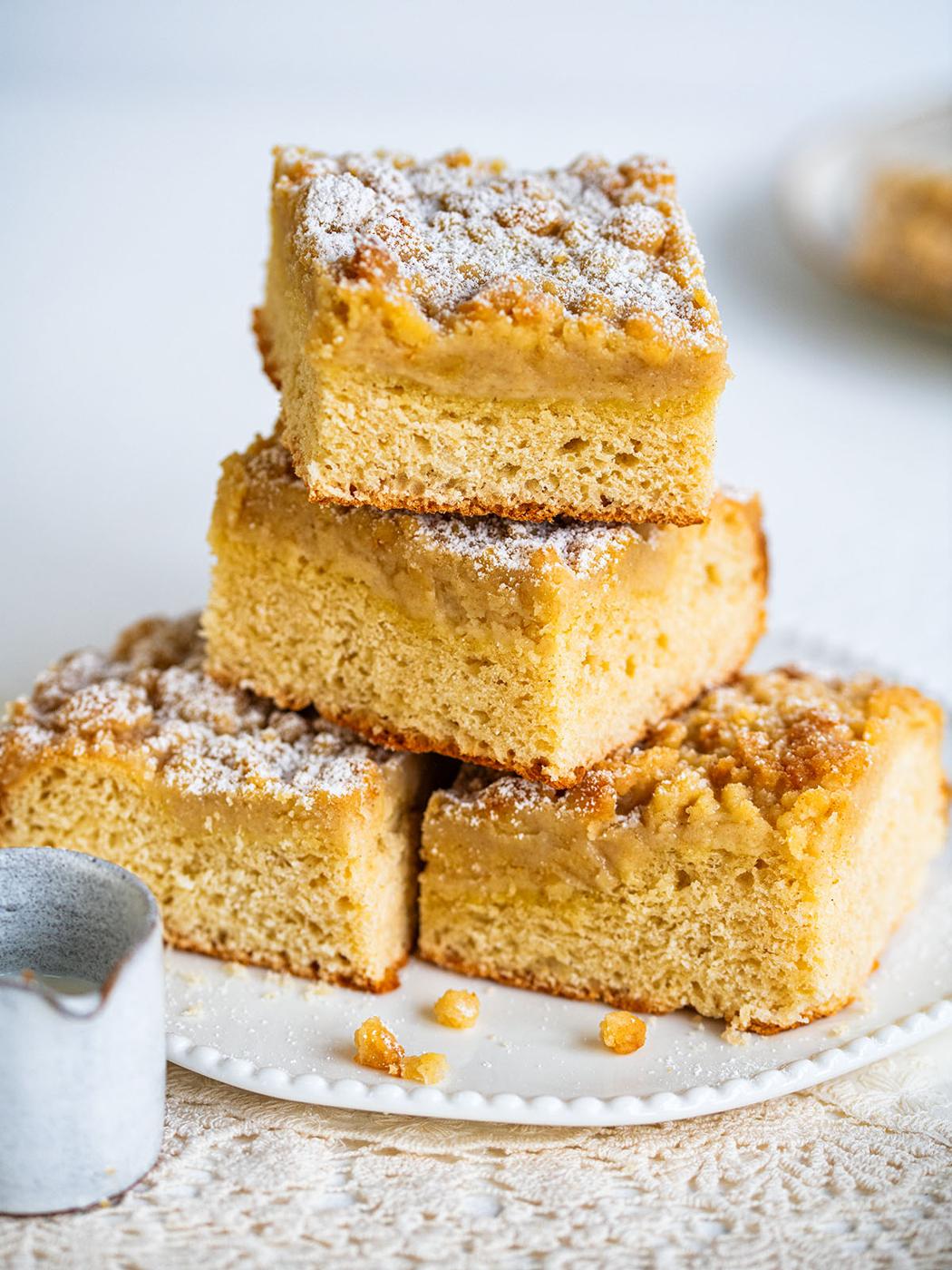 Four square pieces of German crumb cake (streuselkuchen) stacked on a white plate, dusted with powdered sugar. The cakes feature a golden brown crumb layer and a soft, fluffy base. A small pitcher rests nearby on a textured tablecloth.