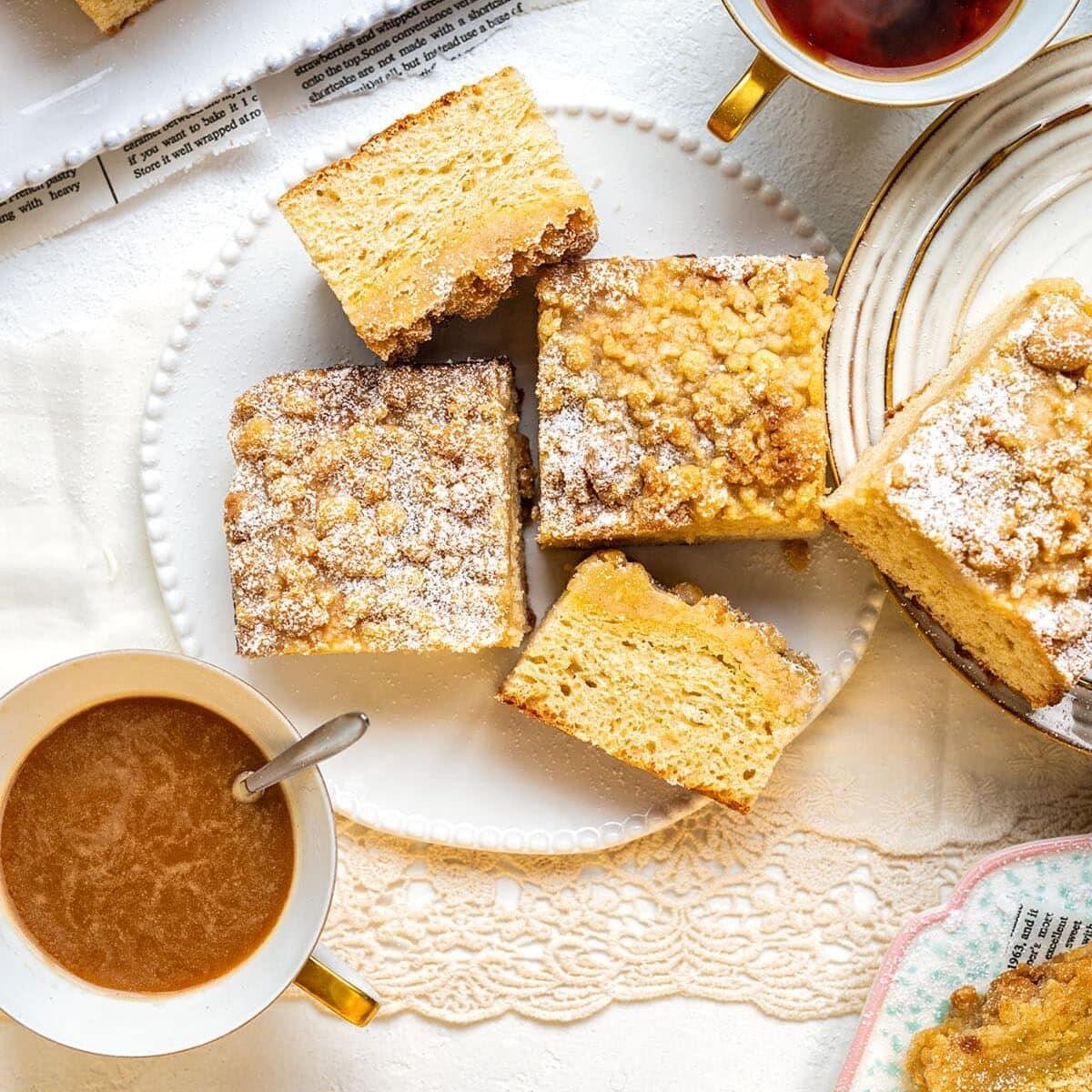 Overhead view of crumb cake squares on a white plate, surrounded by cups of coffee and tea on a lace tablecloth. Some cakes are dusted with powdered sugar and a few crumbs are scattered around.