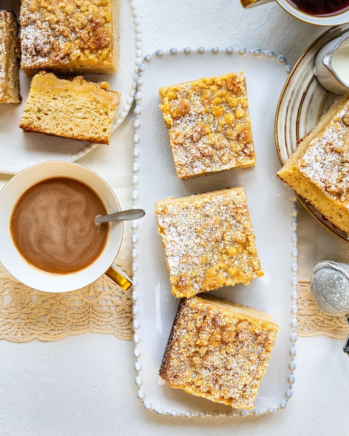 Three slices of crumb cake topped with powdered sugar on a white tray, next to a cup of coffee, a small pitcher of cream, and additional cake slices on a plate, all on a light table setting.