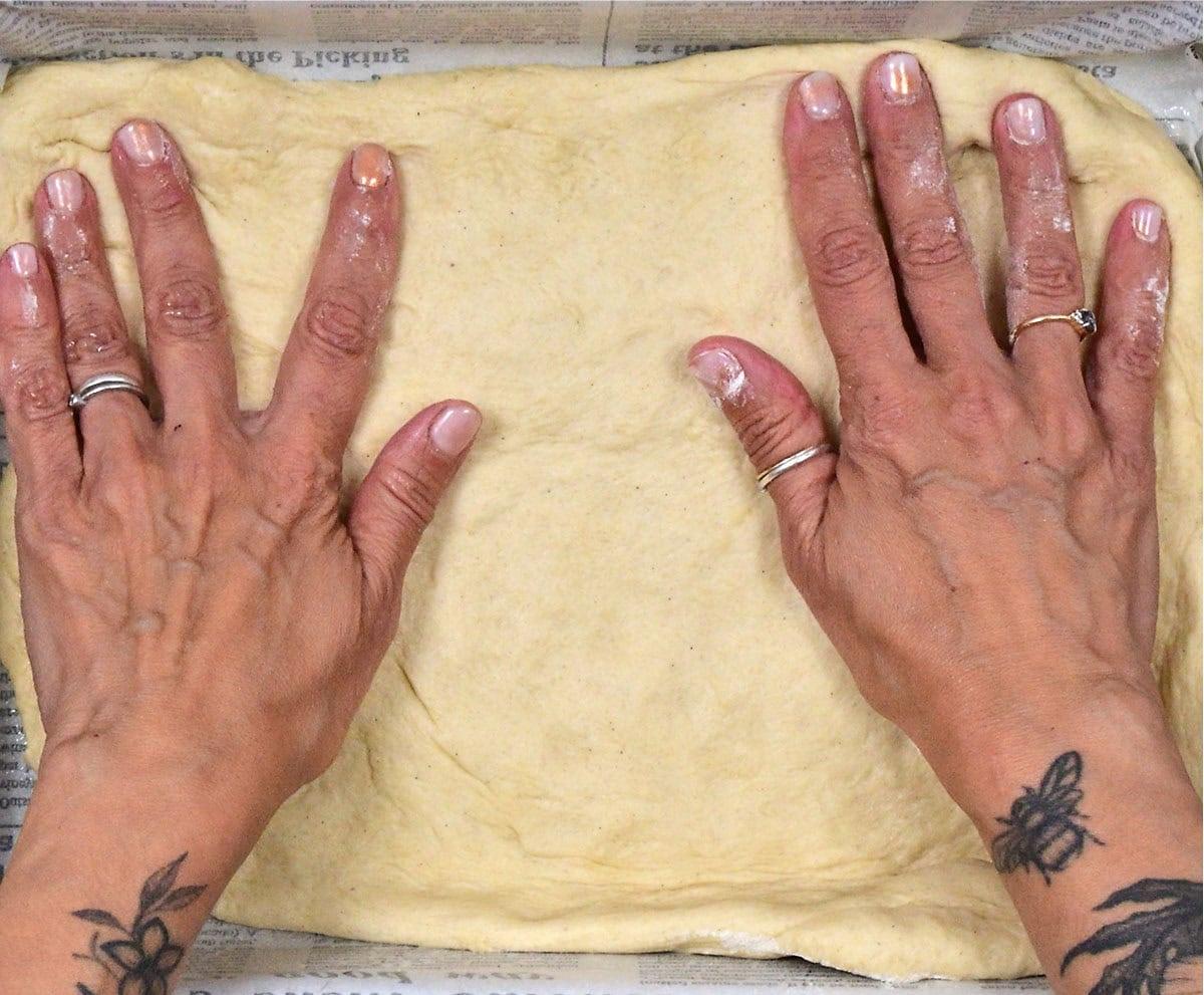 Two tattooed hands with rings press and stretch dough into a rectangular baking pan lined with newspaper. The fingers are spread out, and the nails are painted a light pink color.