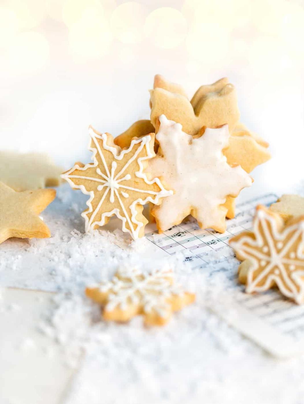 Snowflake-shaped sugar cookies, made from a classic sugar cookie recipe and adorned with white icing, are arranged on a snowy surface with pieces of sheet music in the background, creating a festive and wintry scene.