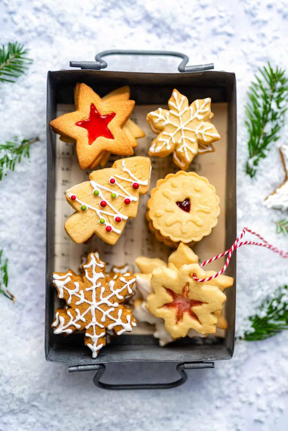 A tray of assorted Christmas cookies, featuring classic shapes like stars and trees from a favorite sugar cookie recipe, is decorated with icing and sprinkles and rests on a snowy surface with pine branches scattered around.