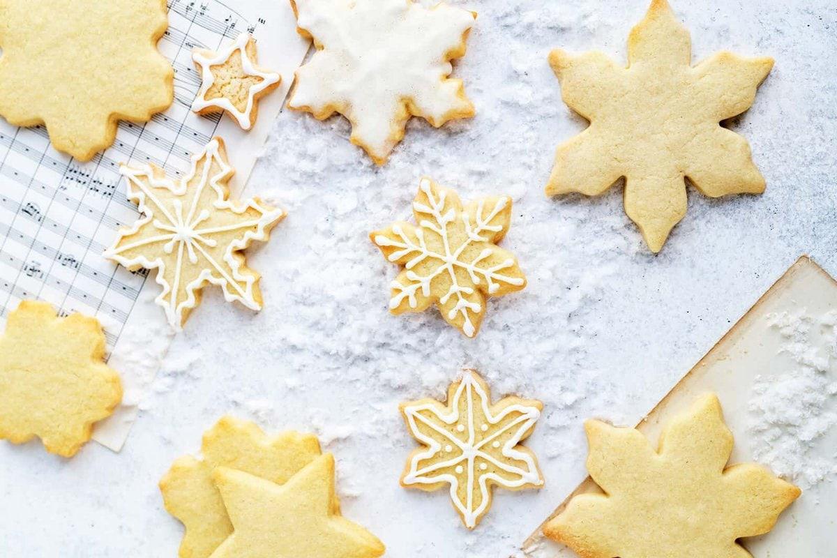 Sugar cookies shaped like snowflakes and stars, some decorated with white icing, are arranged on a floured surface alongside a sheet of music and a wooden board dusted with flour.
