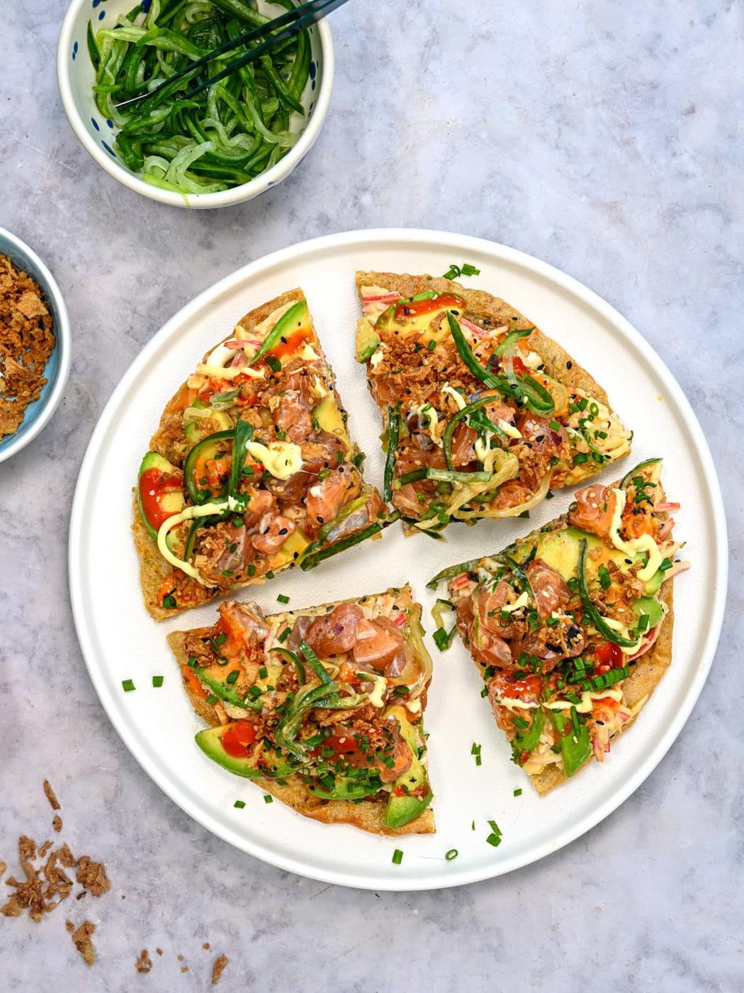 A white plate with four slices of poke-inspired flatbread topped with diced fish, avocado, green onions, sauce, and sesame seeds. Nearby are bowls of sliced cucumbers and crispy onions on a marble surface.