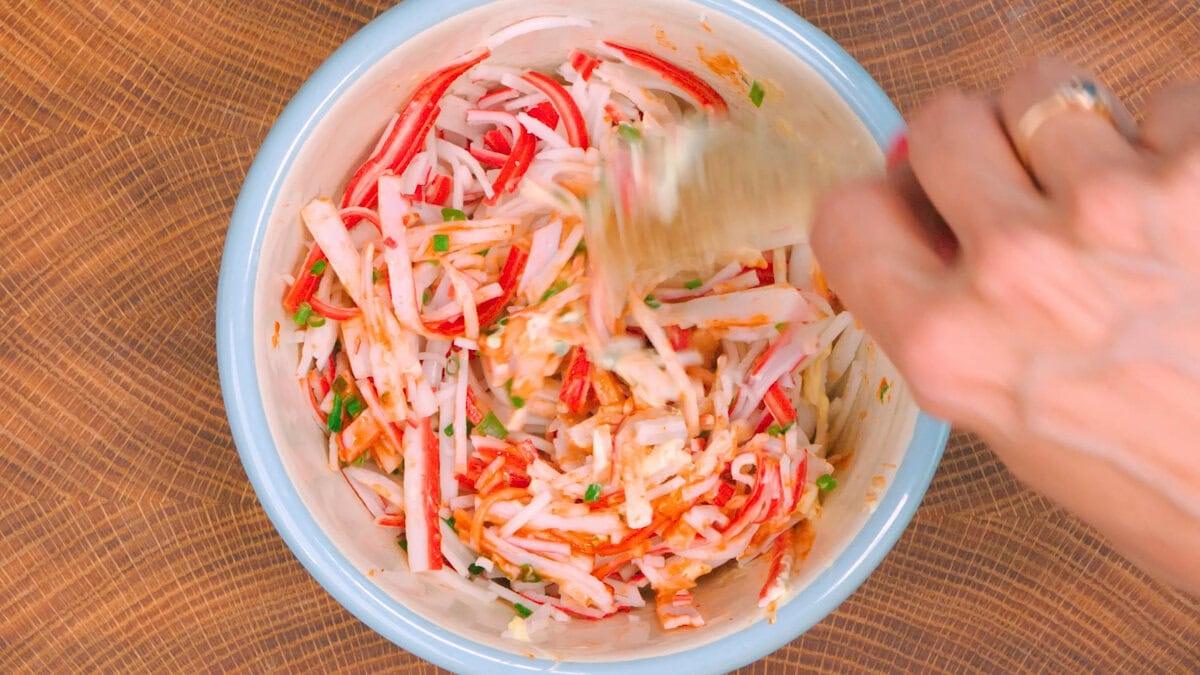 A hand is mixing a salad with imitation crab sticks, thinly sliced vegetables, and herbs in a white bowl on a wooden surface.