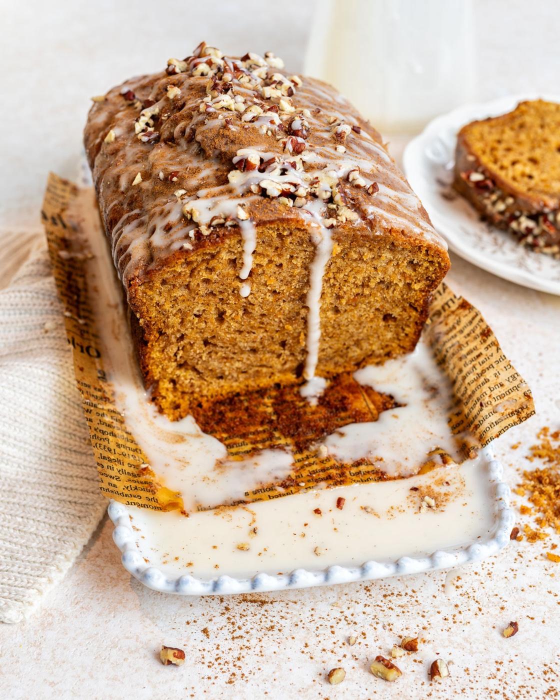 A loaf of glazed sweet potato bread topped with chopped nuts sits on a tray lined with parchment. Glaze drips down the sides, and a slice is cut and placed on a plate in the background. Crumbs and glaze are scattered around.