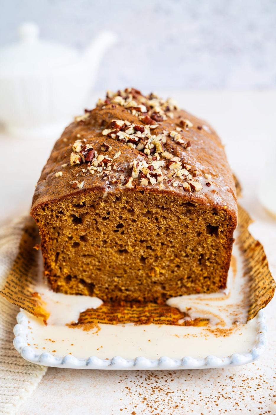 A loaf of glazed sweet potato bread topped with chopped pecans sits on a decorative plate, with a slice missing to show its moist texture. A white teapot and cup are blurred in the background.