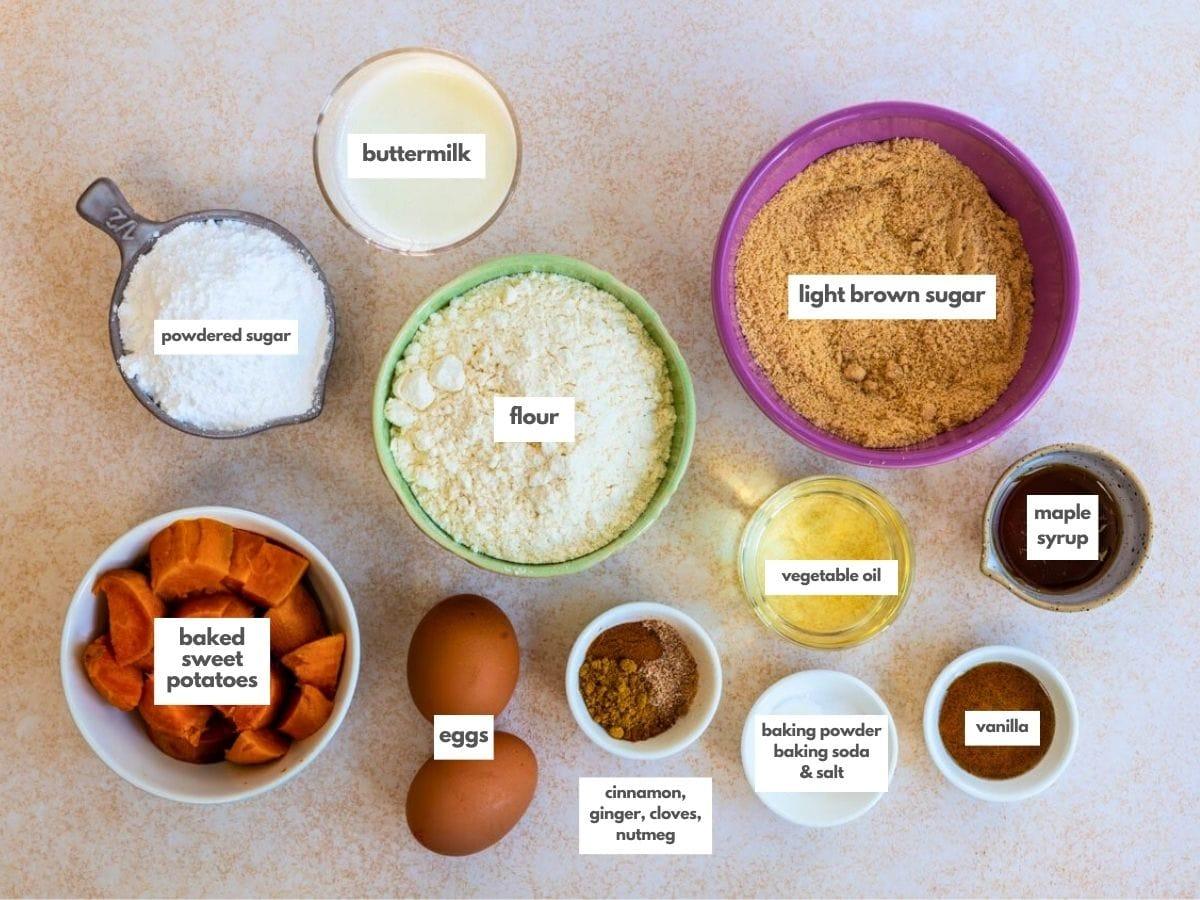 A top-down view of various labeled baking ingredients for Sweet Potato Bread in bowls and cups, including buttermilk, powdered sugar, flour, light brown sugar, maple syrup, baked sweet potatoes, eggs, spices, vegetable oil, and vanilla.