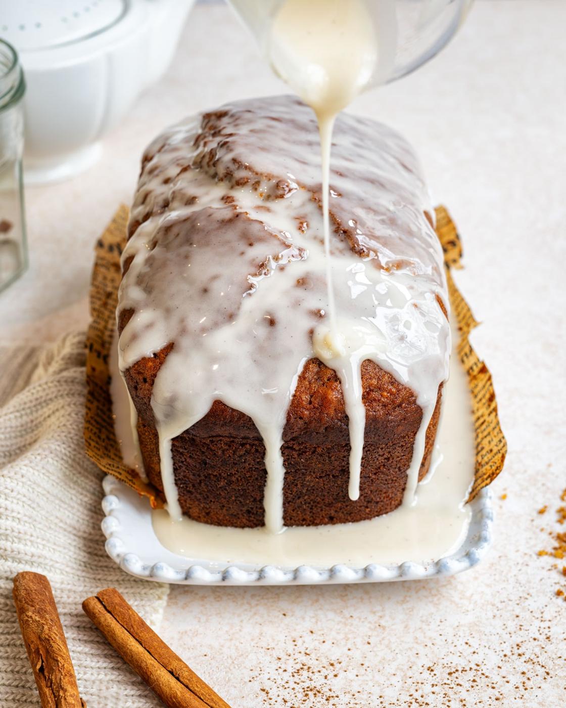A loaf of sweet potato bread sits on a white plate with thick white icing being poured over it. The bread is partially wrapped in parchment paper. Two cinnamon sticks and a cream-colored cloth are beside it.