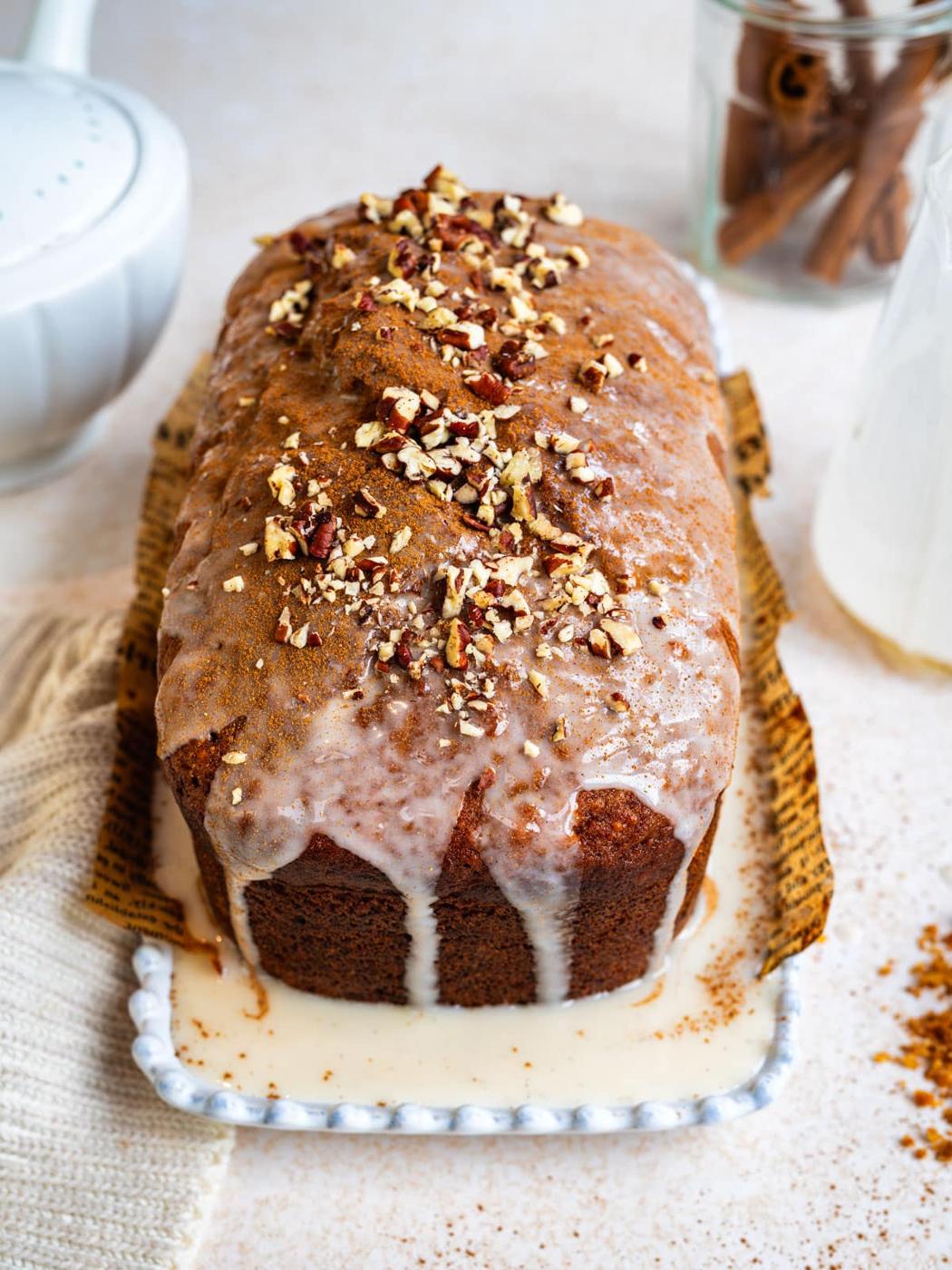 A loaf of glazed bread topped with chopped nuts sits on a white platter. Icing drips down the sides, and cinnamon sticks and a glass bottle are in the background. A piece of parchment is under the bread.