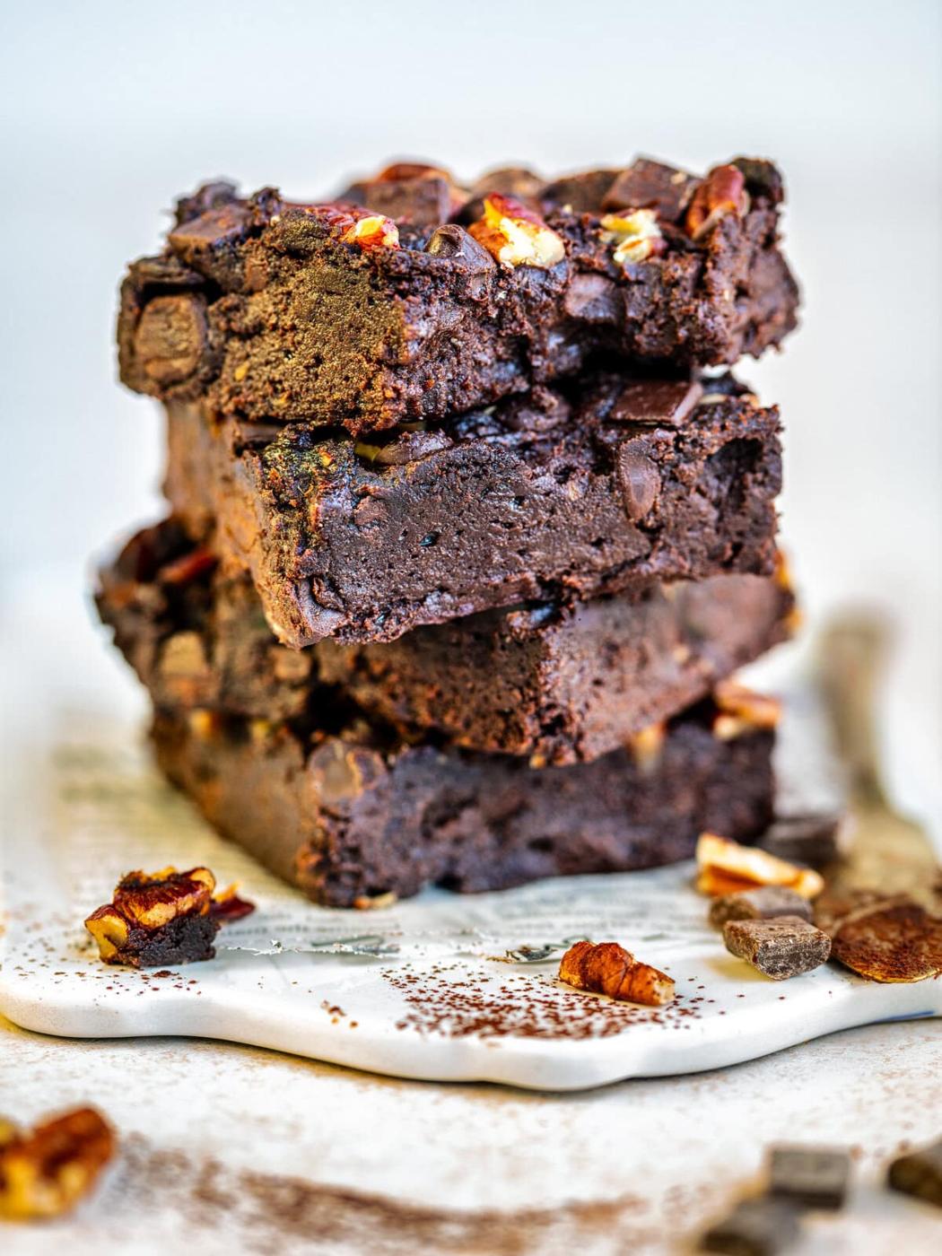 A close-up of a stack of three rich, fudgy chocolate brownies with chocolate chunks and pecans on top, set on a white plate with scattered pecan pieces and chocolate around.