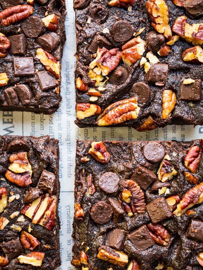 Four sweet potato brownies topped with pecans, chocolate chunks, and chocolate chips are arranged closely together on a sheet of newspaper. The brownies have a rich, fudgy texture and are generously studded with toppings.