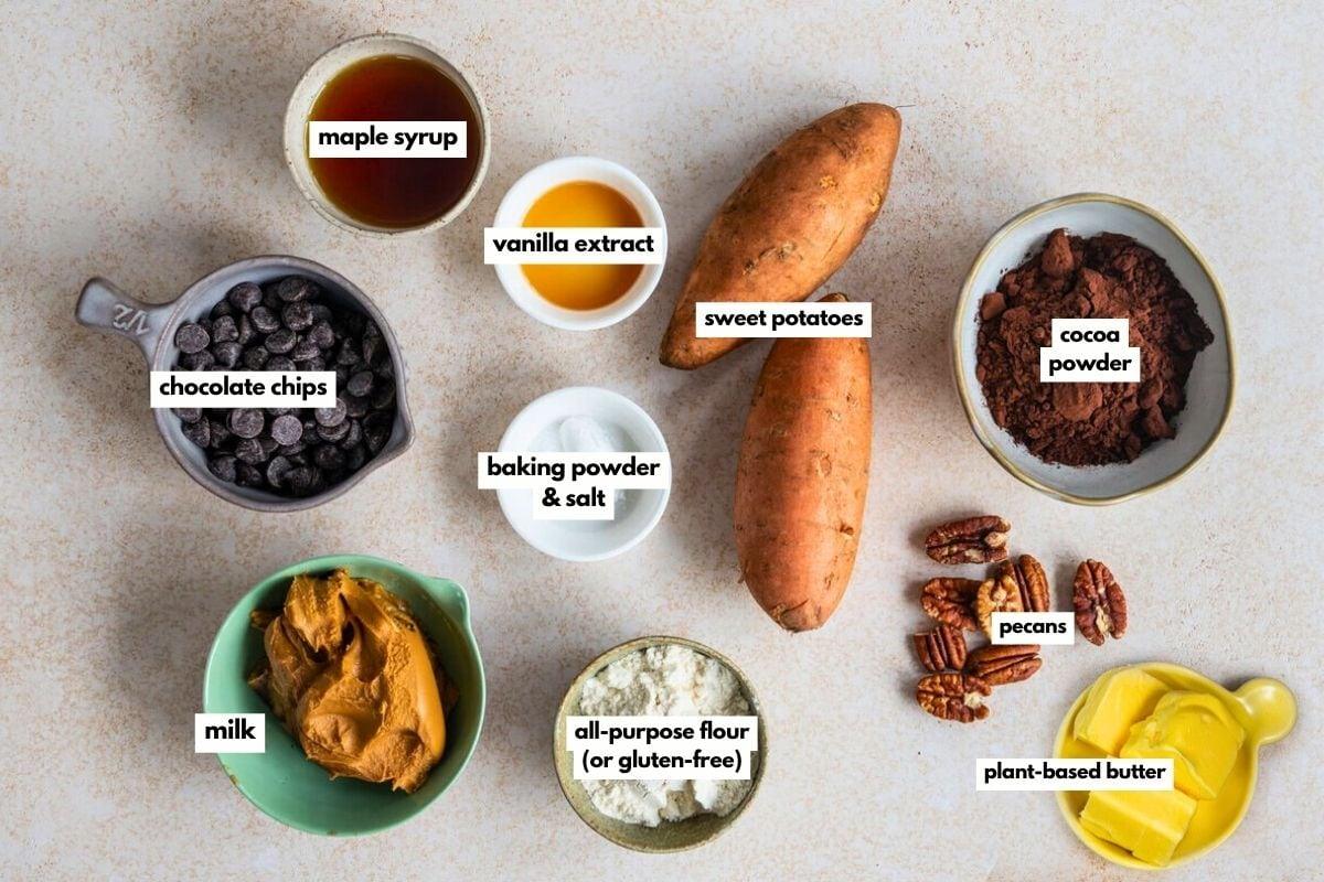 Top-down view of labeled ingredients for vegan sweet potato brownies on a countertop: maple syrup, vanilla extract, sweet potatoes, chocolate chips, cocoa powder, baking powder & salt, pecans, milk, all-purpose flour, peanut butter, and plant-based butter.