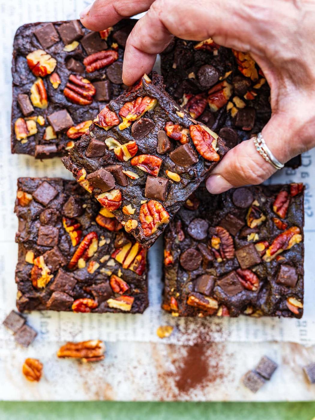 A hand picks up a square of sweet potato brownie topped with pecans and chocolate chunks, with more brownies arranged on newspaper below. Cocoa powder, pecans, and chocolate pieces are scattered nearby.