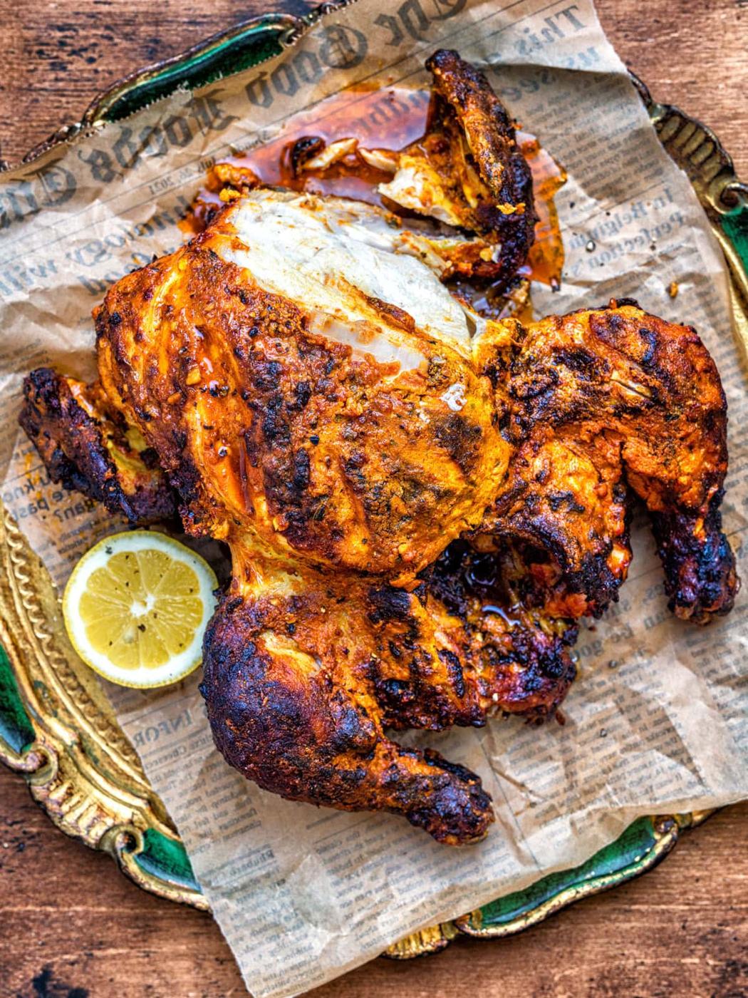 A Tandoori whole chicken with crispy, spiced skin is served on parchment paper with a slice of lemon on the side, presented on a decorative tray.