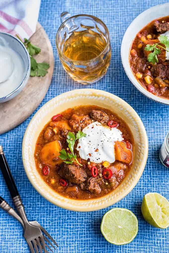 Table set with a yellow bowl of beef stew, limes and beer