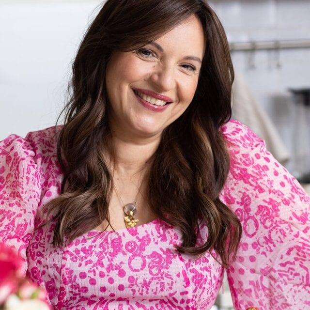 A woman with long brown hair in a pink patterned dress smiles while preparing a dessert with strawberries and peaches in a bright, modern kitchen—perfect inspiration for Thanksgiving recipes. Flowers and fruit are arranged on the countertop.