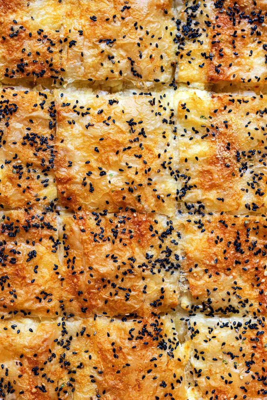 Golden, flaky squares of crisp phyllo pastry, this classic Greek cheese pie—Tiropita—is topped with melted cheese and a sprinkle of black sesame seeds.