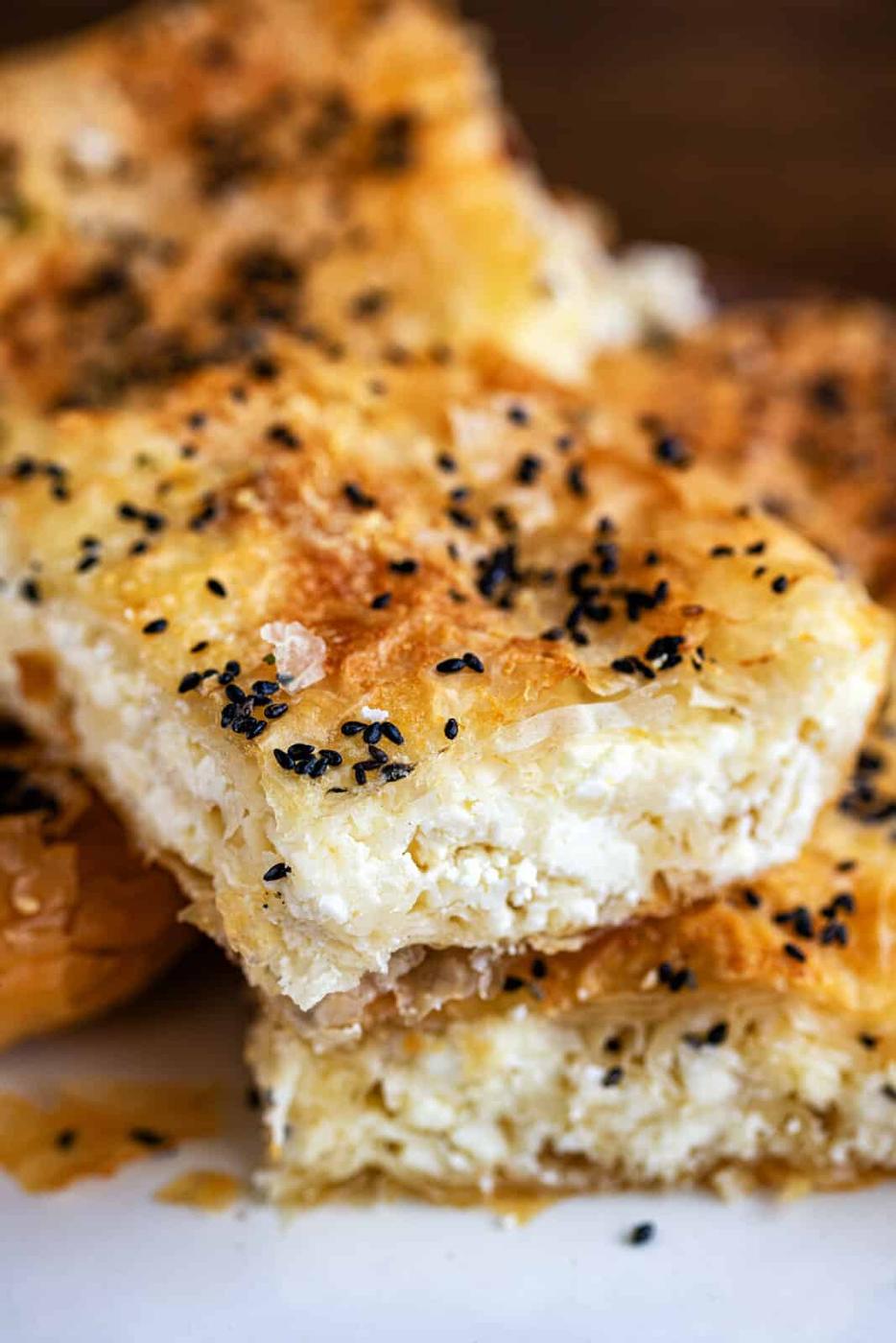 Close-up of flaky, golden-brown Tiropita, with crisp phyllo pastry filled with white cheese and topped with black sesame seeds, stacked on a white surface.