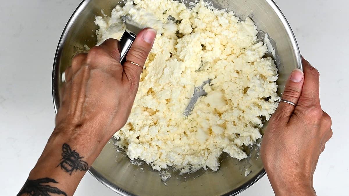 Two hands mix crumbly, pale dough in a large metal bowl—one holds a pastry blender, the other steadies the bowl. A bee tattoo adorns one wrist as they prepare for homemade Tiropita, the classic Greek cheese pie.