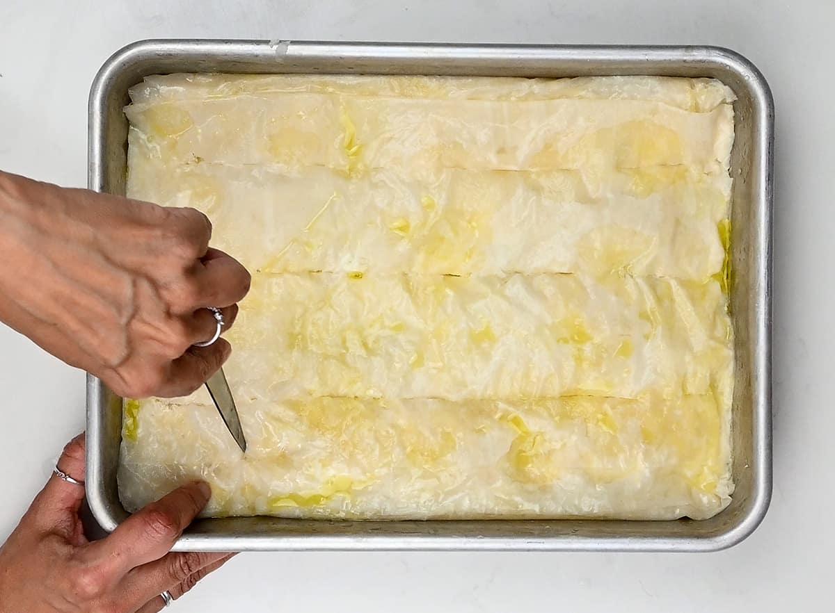 A person is cutting a tray of unbaked tiropita, a Greek cheese pie, with a knife. The crisp phyllo pastry is brushed with melted butter as they hold the tray steady with their other hand.