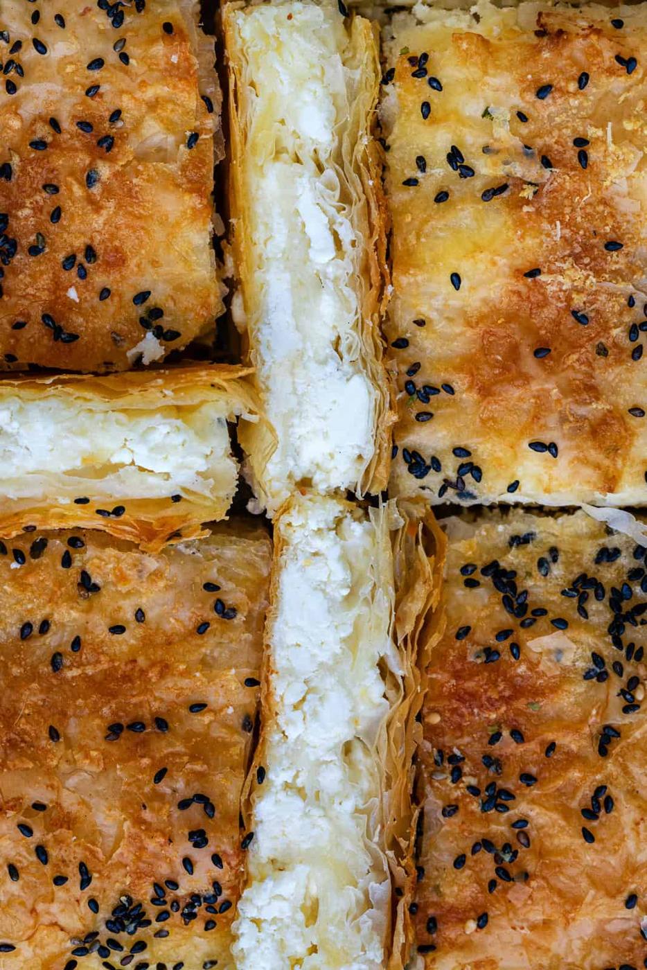 Close-up of golden, flaky Tiropita squares—crisp phyllo pastry filled with creamy white cheese, topped with sesame and black seeds, all arranged closely together.
