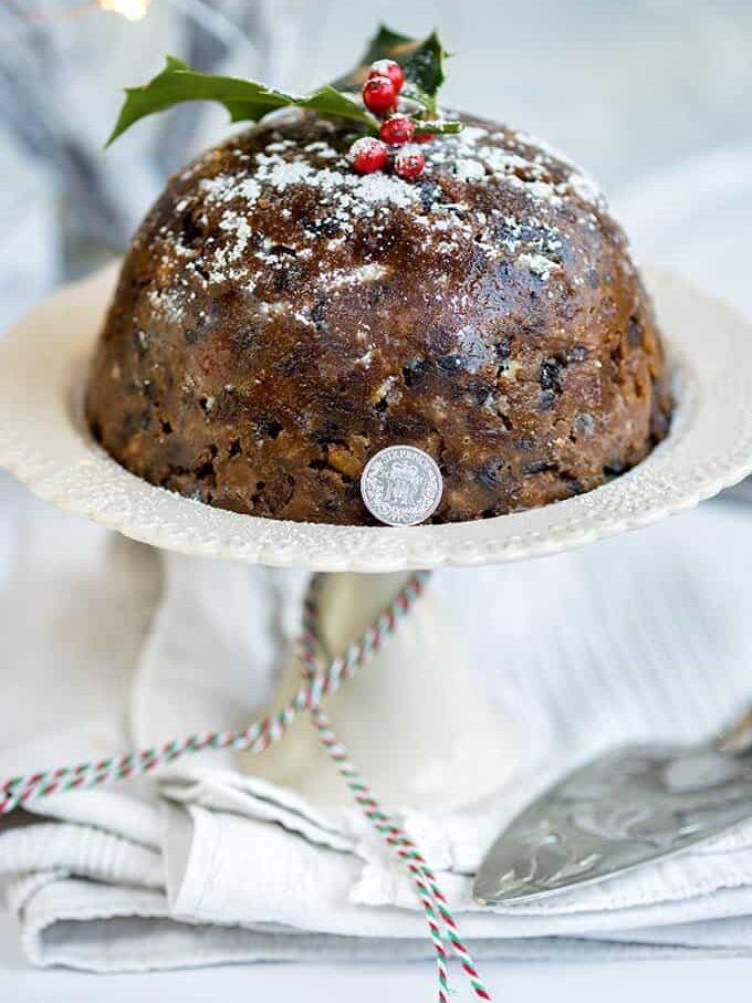 A traditional Christmas pudding decorated with powdered sugar, holly leaves, and red berries sits on a plate. A coin rests against the side of the Christmas pudding, with festive string and a cake server nearby.