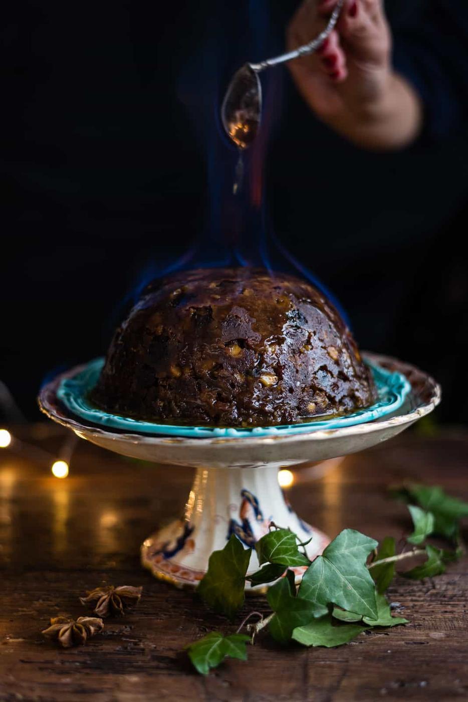 A traditional Christmas pudding blazes with blue flames on a decorative cake stand, while a hand pours sauce over it. Ivy leaves and star anise garnish the rustic wooden table, as warm fairy lights gently glow in the background.